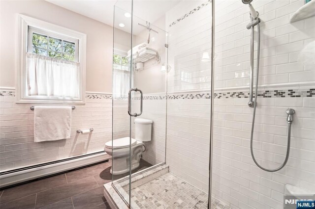 bathroom with a baseboard heating unit, a shower stall, toilet, tile walls, and wainscoting
