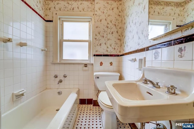 bathroom with wallpapered walls, plenty of natural light, a tub, toilet, and wainscoting