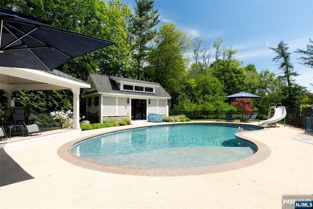 view of swimming pool with a water slide, a patio, an outdoor structure, and view of wooded area