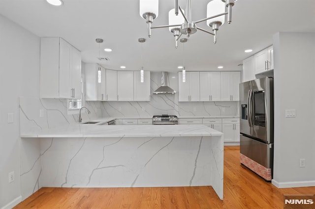 kitchen with stainless steel appliances, wall chimney exhaust hood, a peninsula, light stone counters, and decorative backsplash