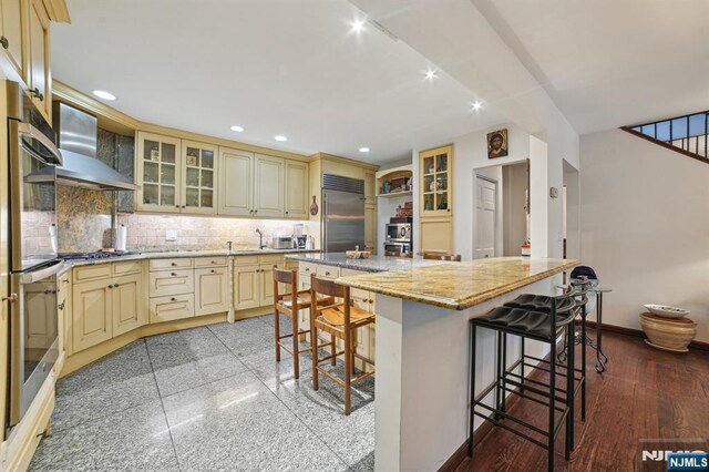 41-21 Dunkerhook Rd, Fair Lawn, NJ 07410 - kitchen with cream cabinetry, stainless steel appliances, wall chimney exhaust hood, recessed lighting, and a kitchen bar