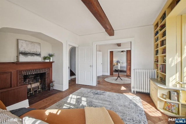 living area featuring radiator heating unit, a brick fireplace, wood finished floors, arched walkways, and beamed ceiling