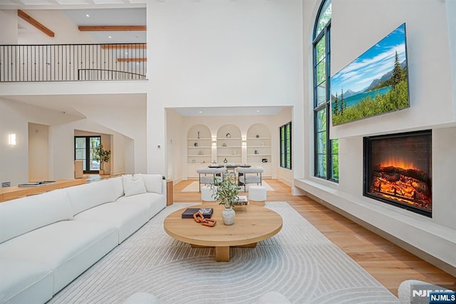 living area featuring a towering ceiling, a warm lit fireplace, light wood-style floors, built in features, and recessed lighting