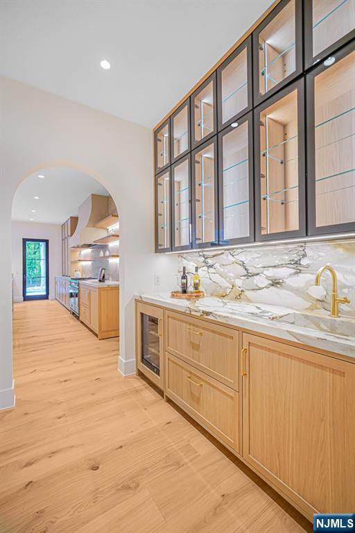 indoor wet bar featuring arched walkways, wall chimney range hood, light wood finished floors, tasteful backsplash, and recessed lighting