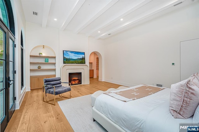 bedroom with light wood-style floors, beam ceiling, arched walkways, a warm lit fireplace, and recessed lighting
