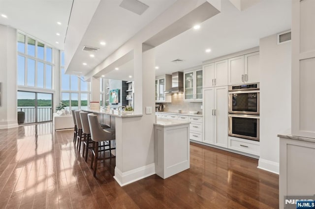 1360 Hamburg Tpke Unit 702, Wayne, NJ 07470 - kitchen with double oven, dark wood-style flooring, wall chimney range hood, glass insert cabinets, and a breakfast bar area