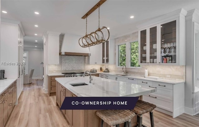 kitchen featuring sink, white cabinetry, light stone counters, custom range hood, and a kitchen island with sink