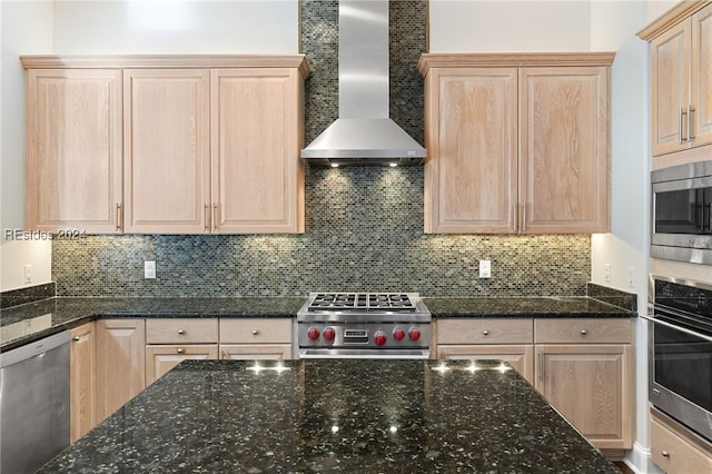 kitchen featuring tasteful backsplash, light brown cabinets, dark stone counters, stainless steel appliances, and wall chimney range hood