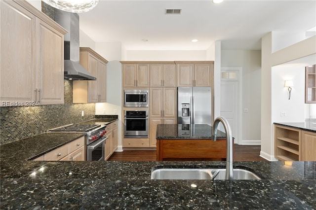 kitchen with appliances with stainless steel finishes, light brown cabinetry, sink, dark stone countertops, and wall chimney range hood