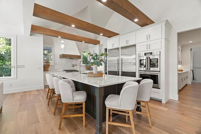 kitchen with a breakfast bar, white cabinets, custom exhaust hood, a large island with sink, and stainless steel appliances