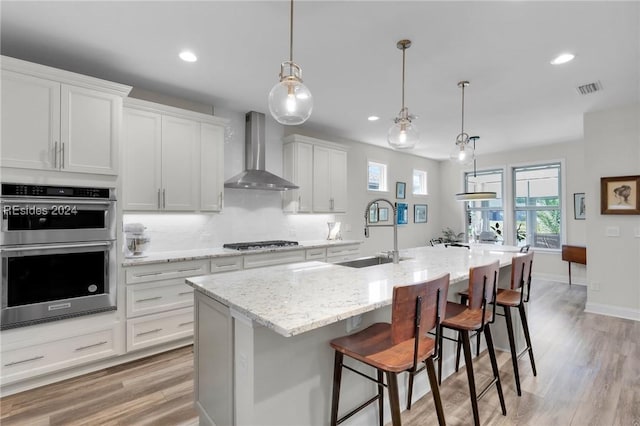 kitchen featuring sink, white cabinetry, stainless steel appliances, a kitchen island with sink, and wall chimney range hood