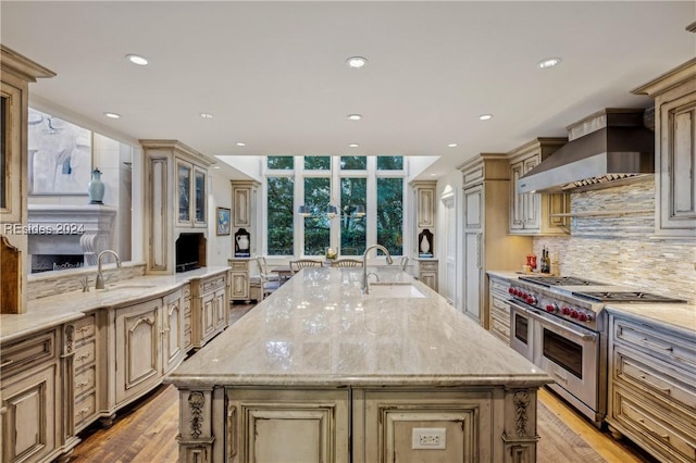 kitchen featuring sink, double oven range, tasteful backsplash, an island with sink, and wall chimney exhaust hood