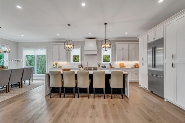 kitchen with pendant lighting, custom range hood, white cabinets, a center island with sink, and built in fridge