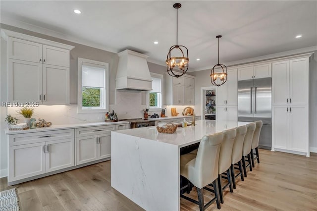 kitchen with custom exhaust hood, white cabinetry, decorative light fixtures, a center island with sink, and appliances with stainless steel finishes