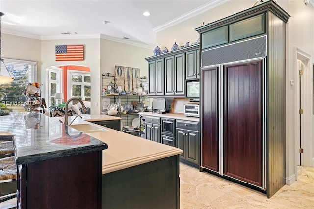 kitchen with sink, hanging light fixtures, ornamental molding, paneled fridge, and an island with sink