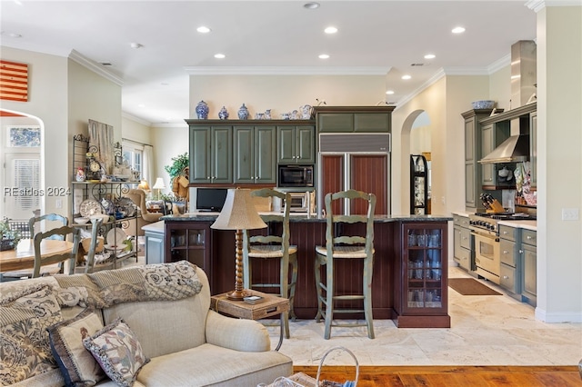 kitchen with a breakfast bar, an island with sink, built in appliances, crown molding, and wall chimney exhaust hood