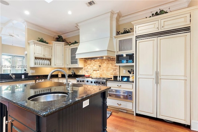 kitchen with sink, custom exhaust hood, built in appliances, dark stone counters, and a kitchen island with sink