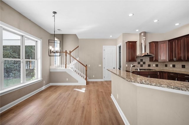 kitchen featuring pendant lighting, decorative backsplash, light stone counters, wall chimney range hood, and light wood-type flooring