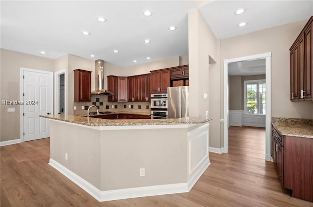 kitchen with wall chimney range hood, appliances with stainless steel finishes, tasteful backsplash, light stone countertops, and light hardwood / wood-style floors