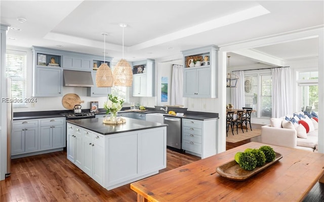 kitchen featuring dark wood-type flooring, dishwasher, hanging light fixtures, a tray ceiling, and wall chimney exhaust hood