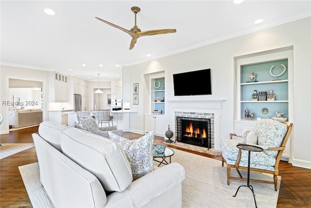 living room with a brick fireplace, built in shelves, dark wood-type flooring, and ceiling fan