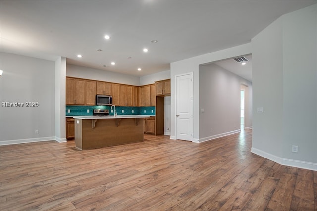 kitchen with light wood-type flooring, a kitchen breakfast bar, an island with sink, stainless steel appliances, and backsplash