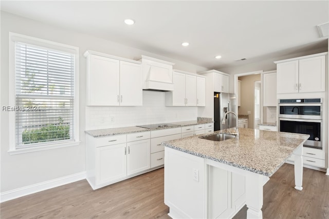 kitchen with sink, white cabinets, stainless steel appliances, custom range hood, and a center island with sink