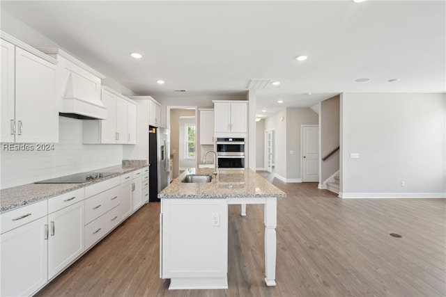 kitchen featuring premium range hood, appliances with stainless steel finishes, sink, a kitchen island with sink, and light stone counters