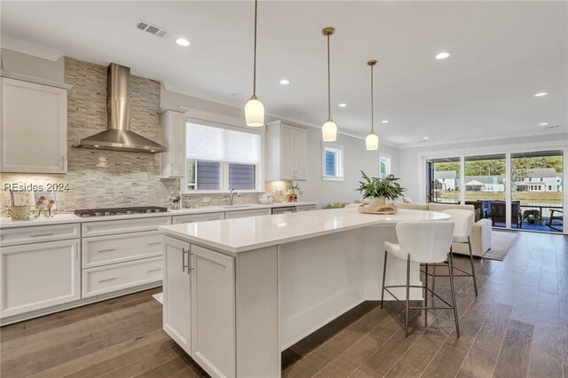 kitchen with white cabinetry, hanging light fixtures, a kitchen island, and wall chimney range hood