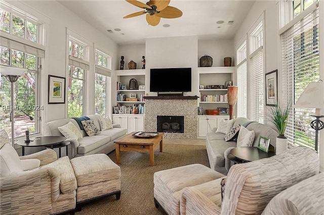 living room with a tiled fireplace, built in features, ceiling fan, and carpet flooring