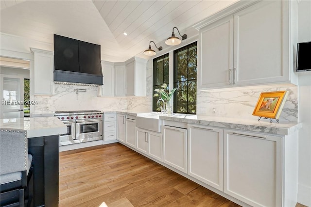 kitchen with vaulted ceiling, white cabinetry, sink, double oven range, and wall chimney range hood