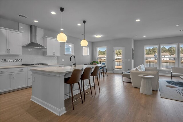 kitchen featuring wall chimney range hood, white cabinetry, hanging light fixtures, tasteful backsplash, and an island with sink