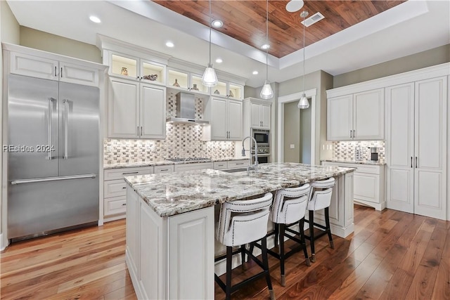 kitchen featuring wall chimney exhaust hood, built in appliances, wooden ceiling, a kitchen island with sink, and white cabinets