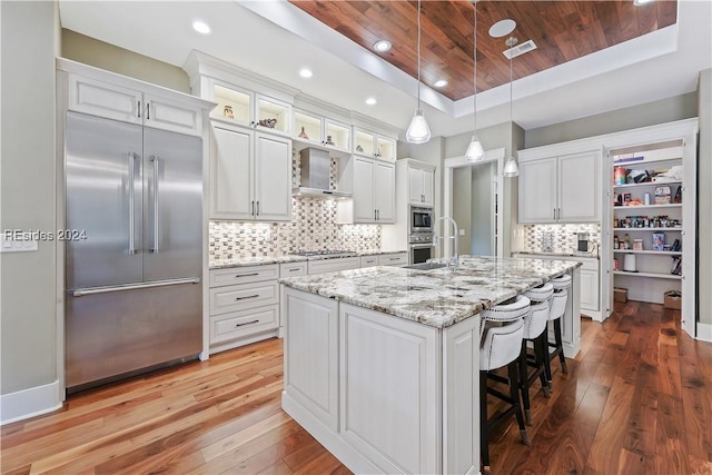 kitchen with white cabinetry, built in appliances, an island with sink, and wooden ceiling