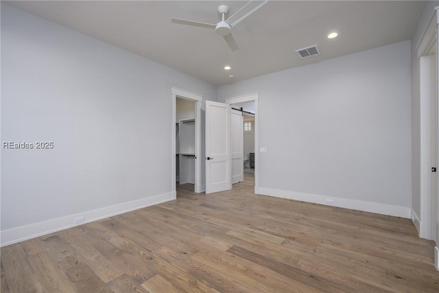 unfurnished bedroom with ceiling fan, a barn door, light hardwood / wood-style floors, and a walk in closet