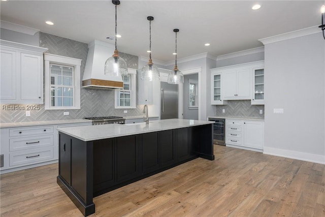 kitchen featuring premium range hood, built in refrigerator, a center island with sink, beverage cooler, and white cabinets