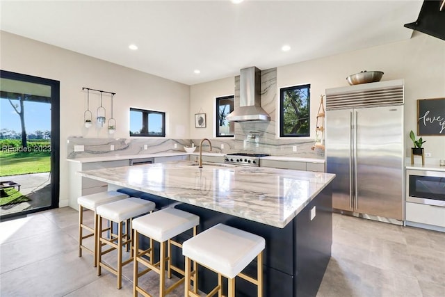 kitchen with light stone counters, wall chimney exhaust hood, built in appliances, and a kitchen island with sink