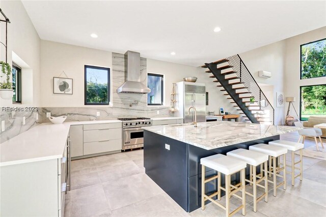 kitchen featuring a breakfast bar area, high end appliances, light stone countertops, white cabinets, and wall chimney exhaust hood