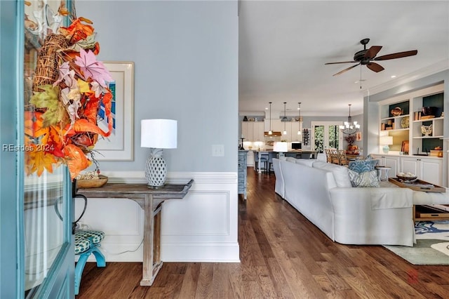 living room featuring dark wood-type flooring, ornamental molding, built in features, and ceiling fan with notable chandelier