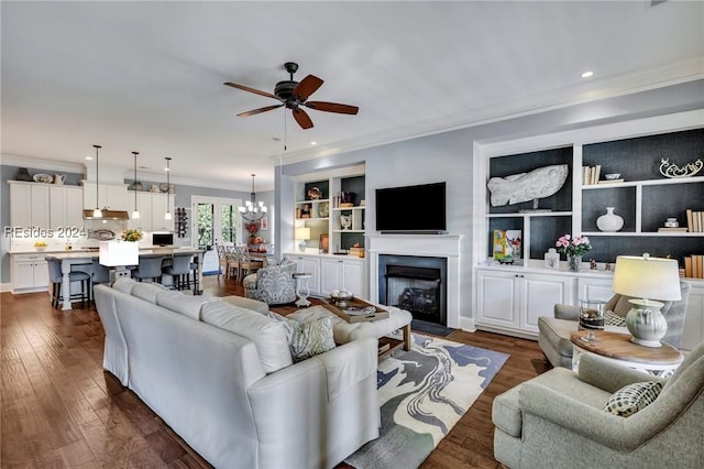 living room with dark wood-type flooring, ornamental molding, ceiling fan with notable chandelier, and built in features