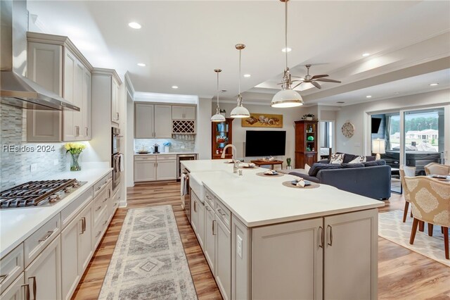 kitchen featuring an island with sink, decorative backsplash, hanging light fixtures, stainless steel appliances, and wall chimney range hood