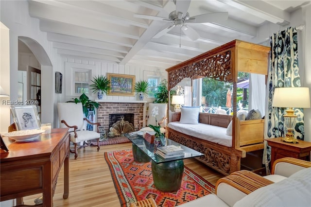 living room with ceiling fan, beam ceiling, a brick fireplace, and light wood-type flooring