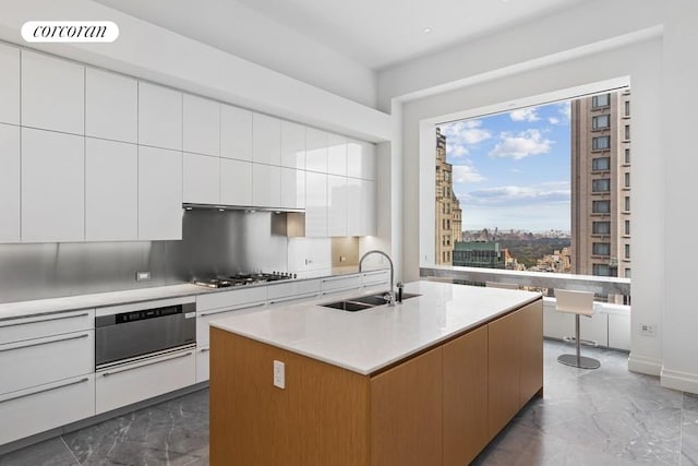 kitchen with decorative backsplash, white cabinets, sink, stainless steel gas cooktop, and a center island with sink