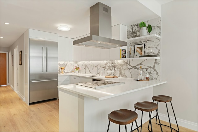 kitchen featuring kitchen peninsula, wall chimney exhaust hood, stainless steel built in refrigerator, a breakfast bar, and white cabinets