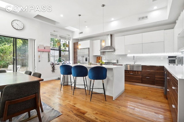 kitchen featuring wall chimney range hood, high end fridge, light hardwood / wood-style floors, tasteful backsplash, and sink