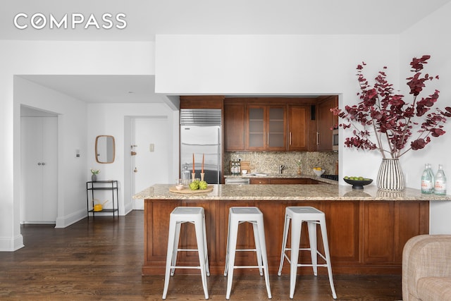 kitchen featuring light stone countertops, dark wood-type flooring, kitchen peninsula, built in refrigerator, and decorative backsplash