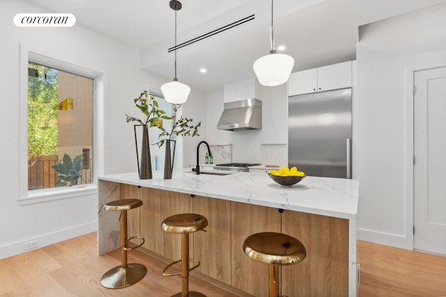 kitchen with light stone counters, white cabinets, hanging light fixtures, wall chimney range hood, and stainless steel appliances