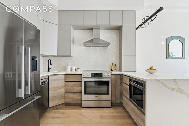 kitchen featuring light wood-type flooring, light stone counters, wall chimney range hood, decorative backsplash, and stainless steel appliances