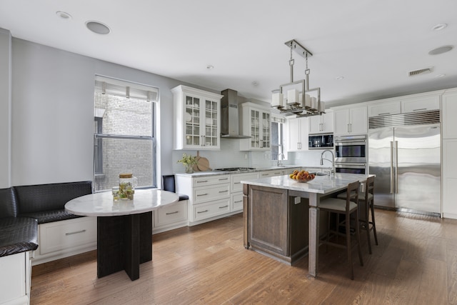 kitchen with a kitchen island with sink, a kitchen bar, built in appliances, wall chimney exhaust hood, and decorative light fixtures