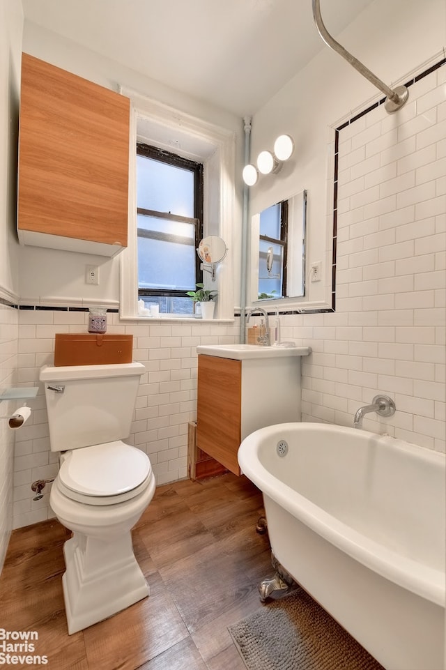 bathroom featuring a tub to relax in, wood-type flooring, toilet, vanity, and tile walls